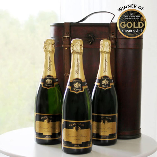 Champagne Triple Celebration Gift Box. Three bottles of champagne are displayed on a round white table. The bottles have gold foil caps and black labels, indicating a premium quality. In the background, a decorative wooden box is partially visible. An award seal in the top right corner reads "WINNER OF MUNDUS VINI GOLD." Natural light illuminates the scene, suggesting an elegant setting.