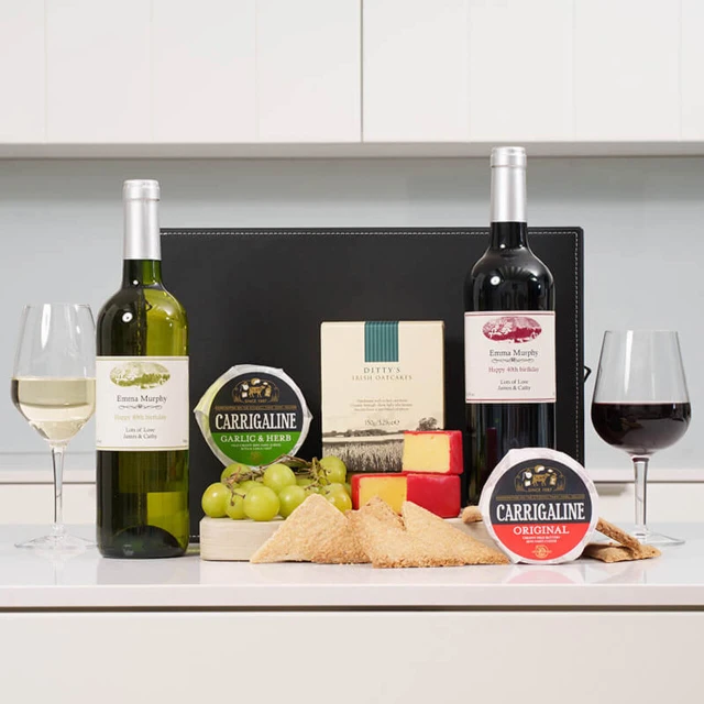 Personalised Fine Wine & Gourmet Cheese Hamper: A selection of two bottles of wine, one white and one red, is displayed alongside two glasses filled with the respective wines. The arrangement includes grapes, various types of cheese, and a box of crackers, all set against a neutral-colored background.