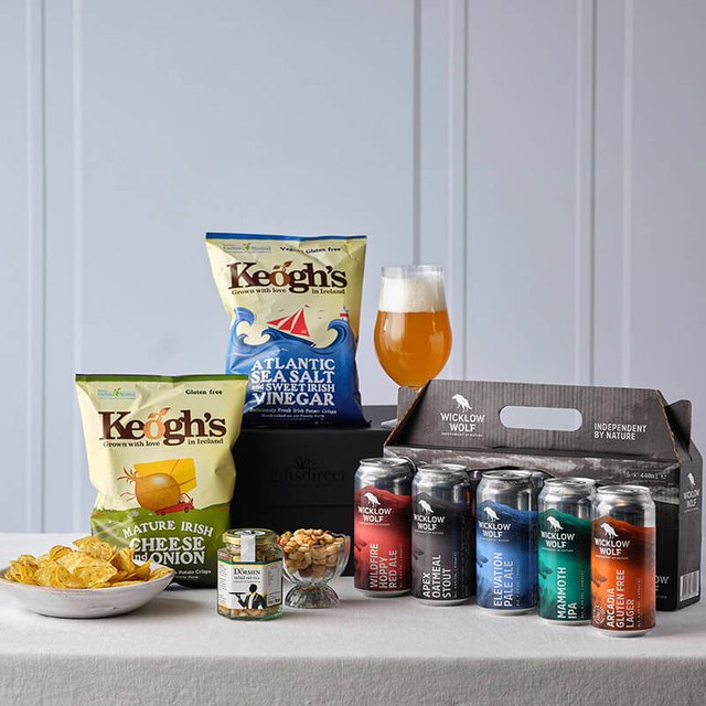 Wicklow Wolf Craft Beer & Snacks Gift Box. A variety of snacks and beverages arranged on a table. There are two bags of Keogh’s potato chips featuring flavors of cheese and onion, as well as Atlantic sea salt and vinegar. A glass of pale-yellow beer sits next to a small bowl of mixed seeds or nuts. In front, a six-pack of assorted Wicklow Wolf beer cans with different colors is displayed inside a black carrier.