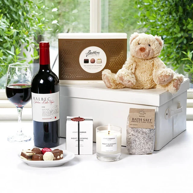 The New Parent Pamper Gift Box. A gift basket arrangement featuring a bottle of red wine, a prop glass of wine, assorted chocolates, a scented candle, bath salts, and a plush teddy bear. The items are presented on a white surface with greenery visible in the background.