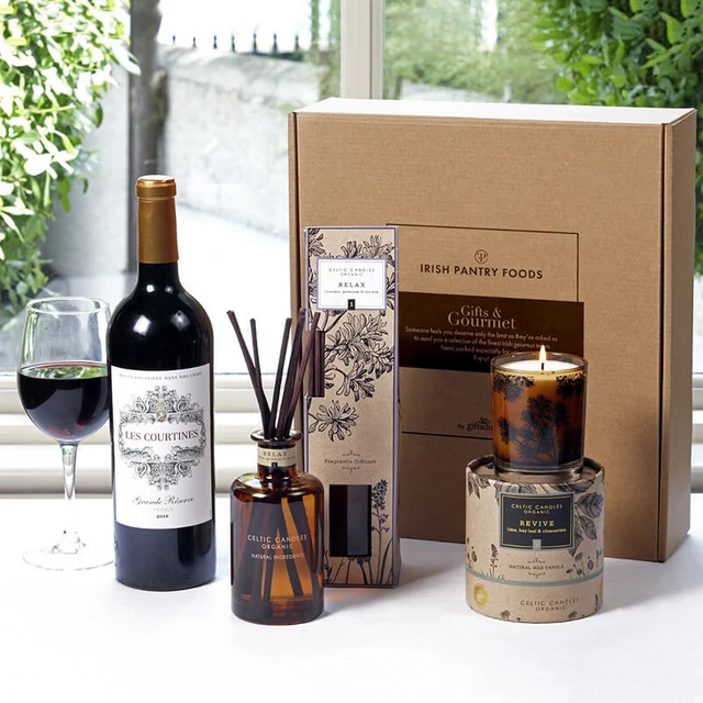 Relax & Revive Pamper Gift Box: A wooden table displays a gift set featuring a bottle of red wine, a scented candle, a diffuser with reeds, and a decorative box labeled "Irish Pantry Foods." A glass of red wine sits next to the bottle.