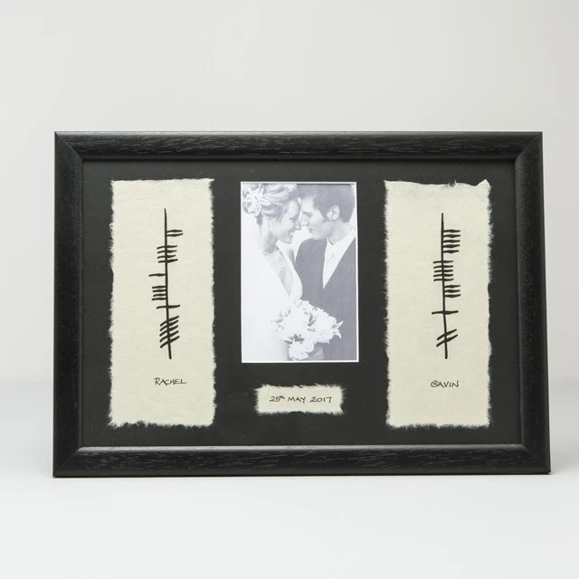 A black-framed photo display featuring a central black-and-white image of a couple on their wedding day, surrounded by two textured white panels. Each panel has Ogham writing and includes the names "Rachel" and "Gavin," along with the date "20th May 2017" on a label at the bottom. The overall aesthetic is elegant and personal.