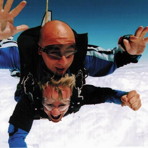 Tandem Skydive Experience Gift. A tandem skydiving experience featuring two individuals in free fall over white clouds. One person, wearing sunglasses and a harness, is positioned above the other, who appears excited with arms raised. The bright blue sky provides a contrasting backdrop to the clouds below.