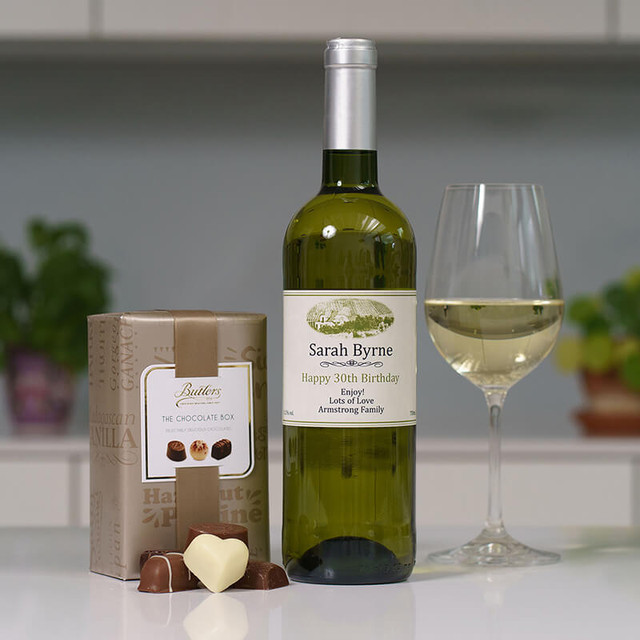Personalised White Wine & 160g Butlers Chocolates. A personalised bottle of white wine labeled "Sarah Byrne" next to a glass filled with the same wine. A box of chocolates is placed nearby, with one chocolate shaped like a heart in front. The background suggests an indoor setting with plants visible.