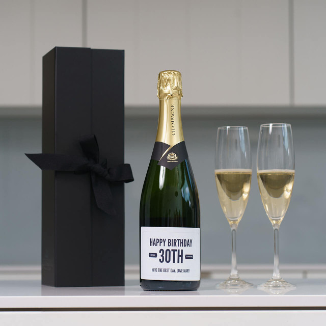 Personalised champagne bottle labeled ‘Happy 30th Birthday – Have the Best Day Love Mum,’ displayed with two filled flutes and a black ribboned gift box on a white surface.