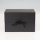 A closed black box with a ribbon tie, sitting on a white surface. The box has a minimalist design and features a logo on the top. A closed black box with a ribbon tie, sitting on a white surface. The box has a minimalist design and features a logo on the top.