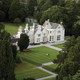 Lough Rynn Castle Leitrim 2 Night Break 1 Evening Meal for 2. Aerial view of a large, historic stone mansion surrounded by lush greenery. Well-maintained lawns and pathways lead to the entrance. Two people walk on a path in the foreground. Lough Rynn Castle Leitrim 2 Night Break 1 Evening Meal for 2. Aerial view of a large, historic stone mansion surrounded by lush greenery. Well-maintained lawns and pathways lead to the entrance. Two people walk on a path in the foreground.