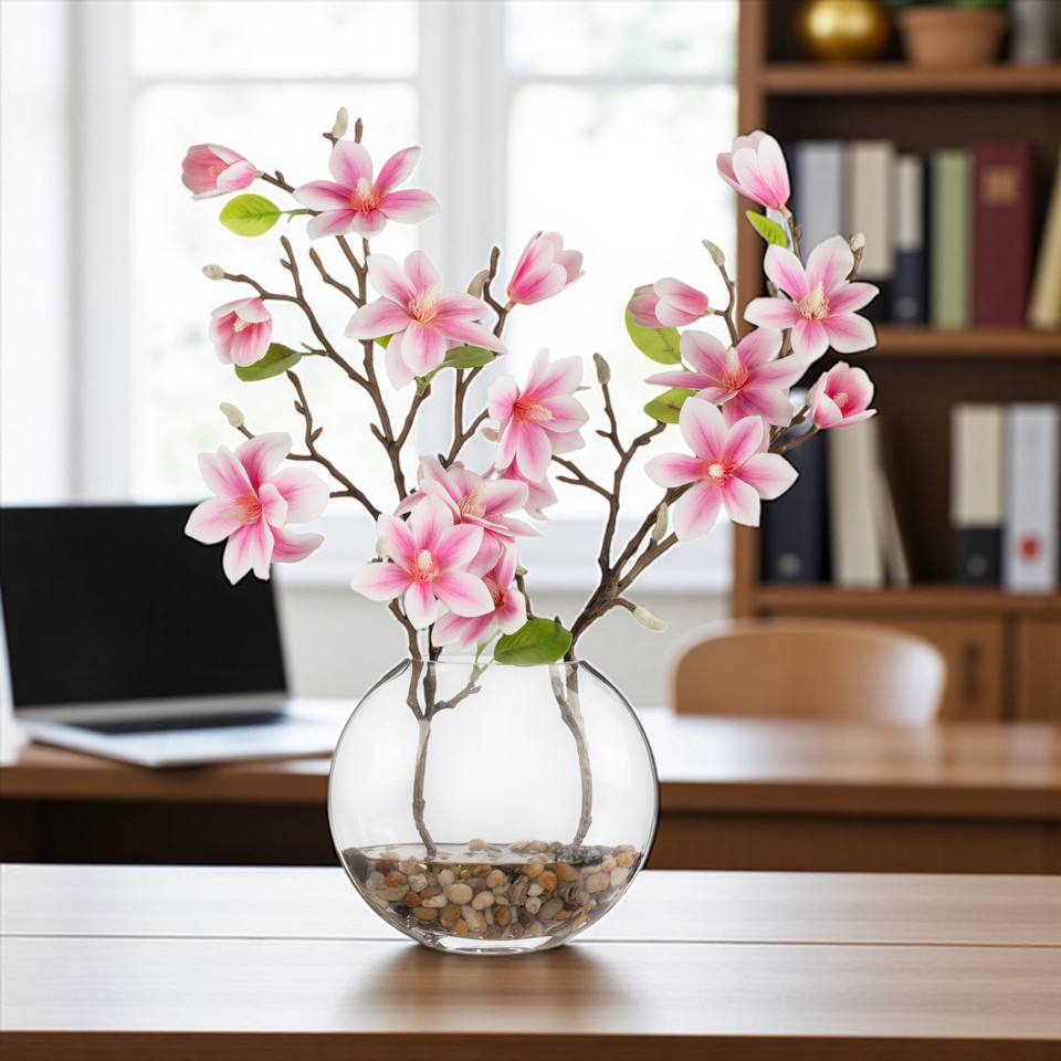 Pink real touch artificial magnolia flower arrangement in a clear round glass vase with faux water and river rock, styled on a desk in a bright home office setting.