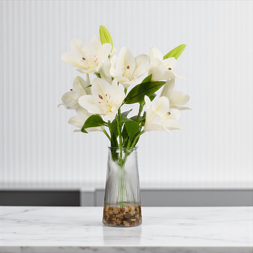 White real touch artificial tiger lily arrangement with green buds displayed in a clear glass vase with faux water, photographed on a tabletop with a neutral modern backdrop.