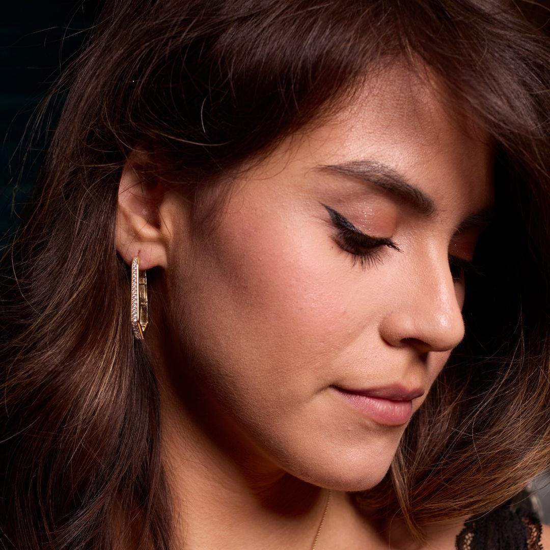 A profile view of a 14K yellow gold oval huggie hoop, showcasing the thin band and the all-around sparkle on a marble background.