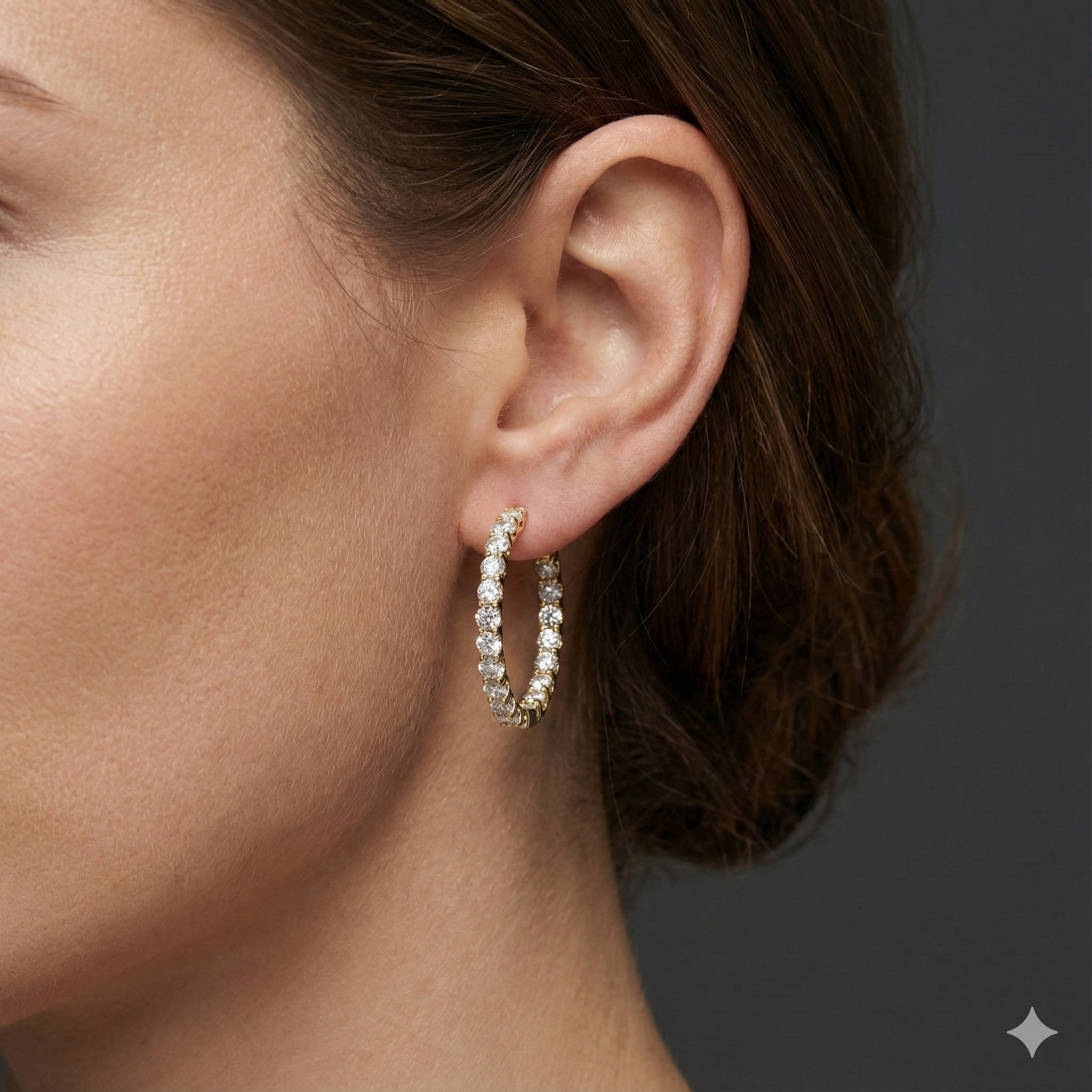 A close-up of a pair of 14K yellow gold huggie hoop earrings with a continuous line of brilliant round-cut diamonds, shown on a black background to highlight their sparkle on ear.