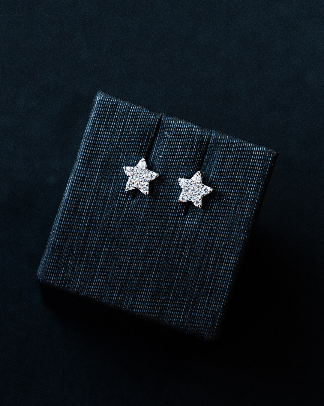 A pair of sparkling, star-shaped stud earrings displayed on a dark, textured jewelry box insert against a black background.