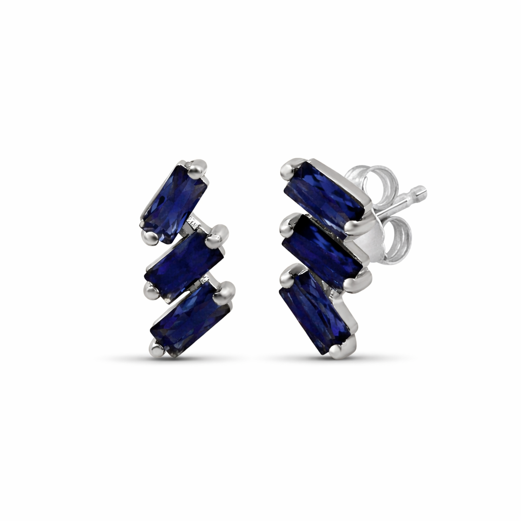 A pair of cascading stud earrings featuring a vertical arrangement of three baguette-cut blue sapphires. The rectangular gemstones are staggered in a step-like formation and held in a minimalist 18k white gold setting, shown against a clean, white studio background.