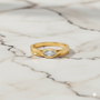 A top-down view of a 14K yellow gold bypass ring with a single, horizontally-set oval diamond in a bezel setting on a marble background.