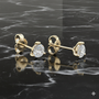 A pair of 14K yellow gold three-prong diamond stud earrings, shown from a side profile on marble.