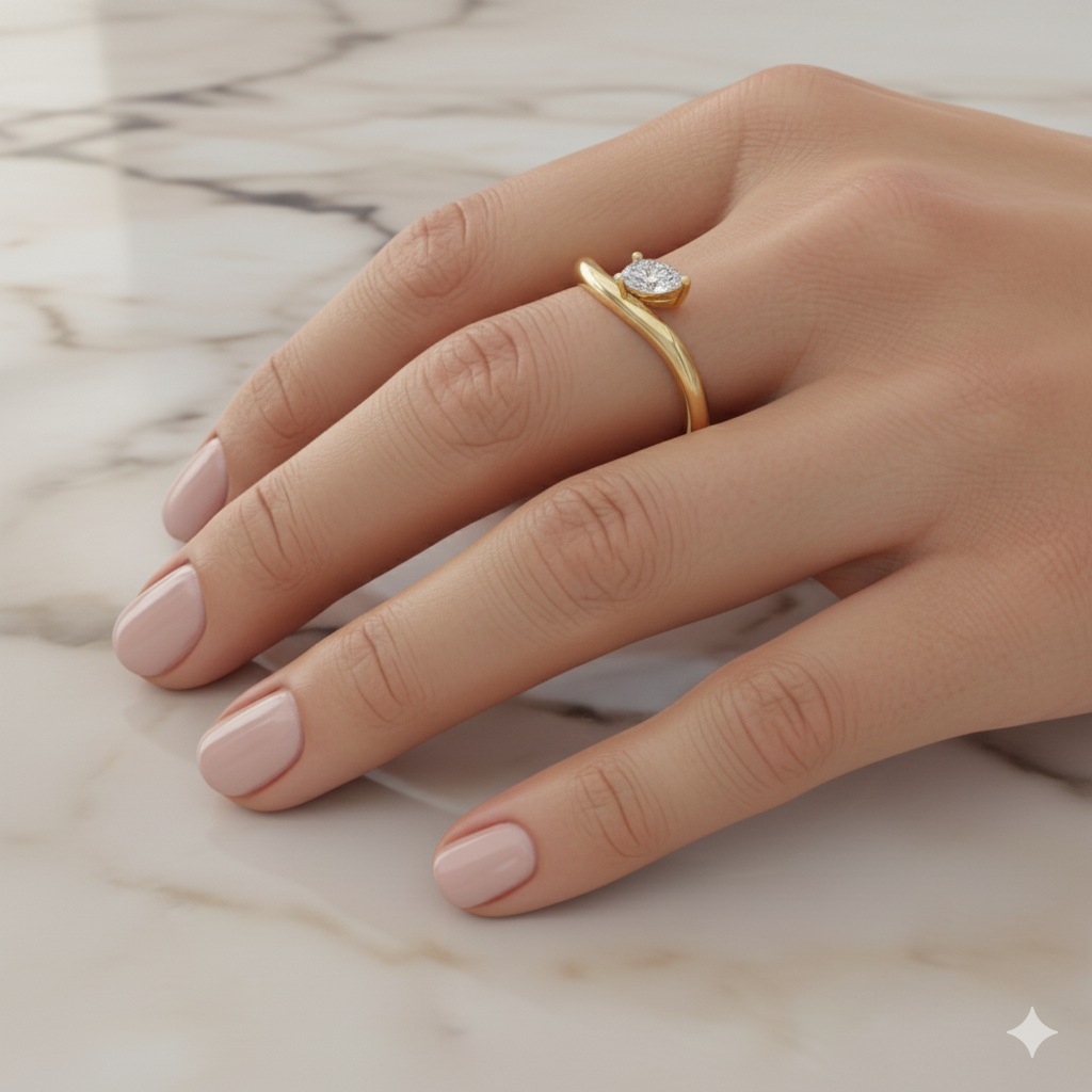 A view of a 14K yellow gold bypass ring with a single pear-shaped diamond at one end of a fluid, curving band, on a finger of a hand.