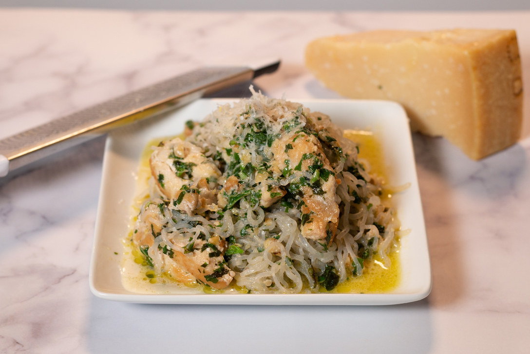 A white square plate of It's Skinny angel hair pasta with chicken cooked creamy pesto sauce. The plate sits next to to a block of parmesan cheese.