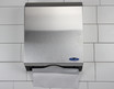 Wall-mounted Frost 105 multifold paper towel dispenser in Stainless Steel, installed on a tiled wall with a partially visible paper towel protruding from the bottom.