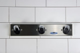 A Frost 1150-3 Safety Coat Hook Strip mounted on a tiled wall, demonstrating its durable construction and safe, triple-hook design for efficient use in any professional environment.