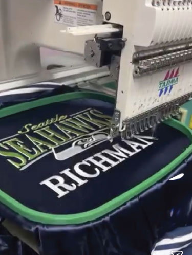 A multi-needle embroidery machine stitching a team name and player name onto a navy jersey held in a hoop.