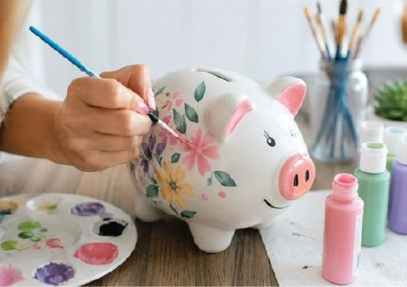 A hand painting delicate flowers onto a white ceramic keepsake bank, surrounded by paint bottles and a palette.