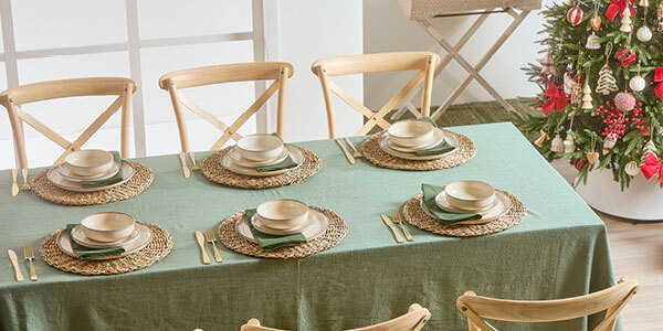 A table with place settings and a green table cloth sits in the middle of the image with chairs around it. In the right corner of the room is a Christmas tree. 