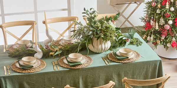 A table is set with six places, in the middle a Christmas garland, candle holders and vase with greenery create a centrepiece. In the right hand side of the room is a decorated Christmas tree. 