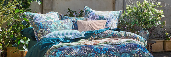 Picture features a bed with botanical print bedding styled with cushions and a throw. Around the bed are styled artificial plants and bouquets. 