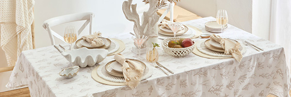 Picture features a set table with a coastal theme. Table cloth features coral designs and the table is set with natural and white coloured placemats, natural coloured tableware, clear and white striped glasses and coral and shell shaped decor pieces. 