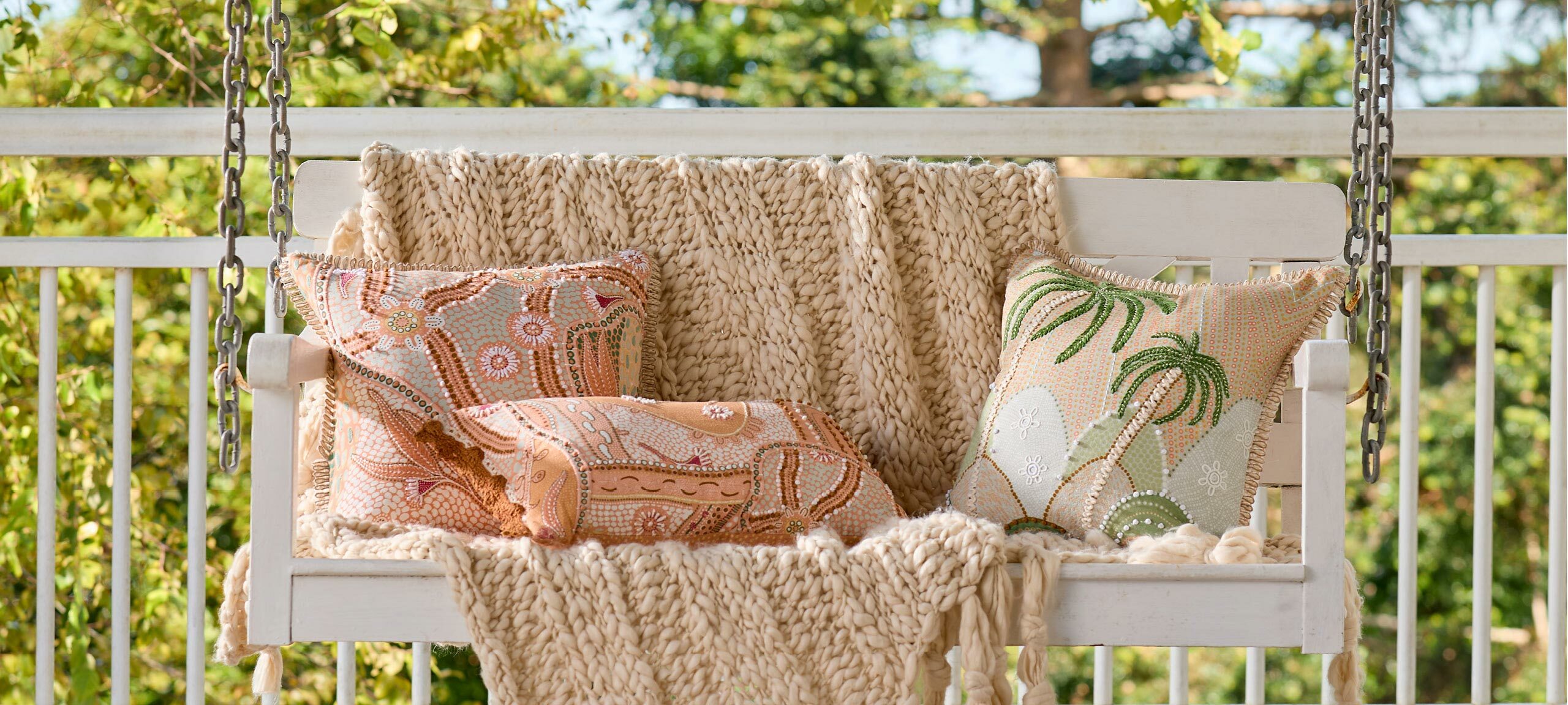 Three cushions, two square and one oblong, and a decorative throw sit on a white swinging chair on a porch. One of the square cushions features an embroidered palm tree design. The other square cushion and the oblong cushion feature an Indigenous Australian design in pink and orange tones, both with textural details. 