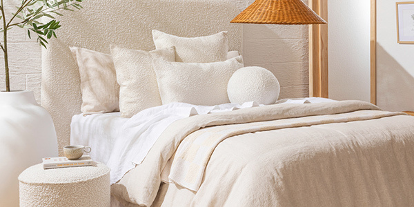A styled bed with cream boucle cushions and natural bedding. Bed features a boucle bedhead and next to the bed is a boucle ottoman and an artificial olive tree in a white pot. 