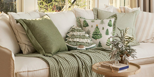 A frosted mini Christmas tree sits on a table in front of a couch. Couch features natural and white cushions with Christmas themed cushions in the middle. 