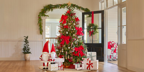 A traditional large green Christmas tree sits in the middle of the room, decorated with red, white and green decorations. Next to the tree sit presents and gnomes decorations that are wearing santa outfits.