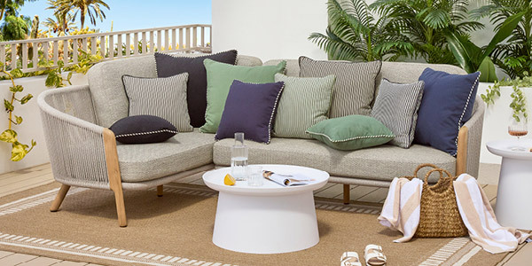 An outdoor couch decorated with Pillow Talk cushions. Underneath the couch is an outdoor rug and in front of the couch is a white outdoor coffee table, set for entertaining with glasses, a carafe and a magazine. 