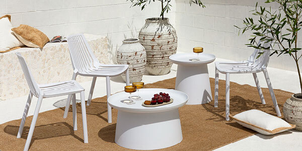 Outdoor dining chairs are set up around an outdoor coffee and outdoor side table, with an outdoor rug underneath them. The tables have glasses and a fruit platter on them. 