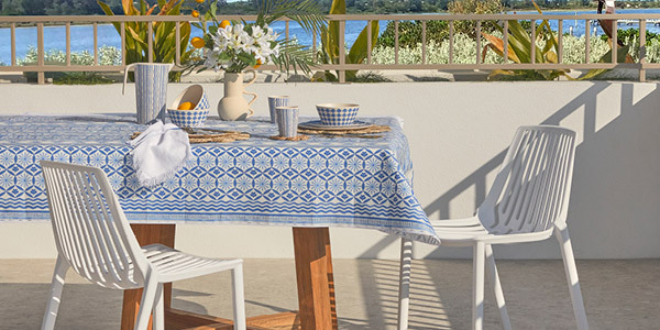 A table is set with an outdoor table cloth, outdoor tableware and dinnerware with two white outdoor dining chairs next to the table. 