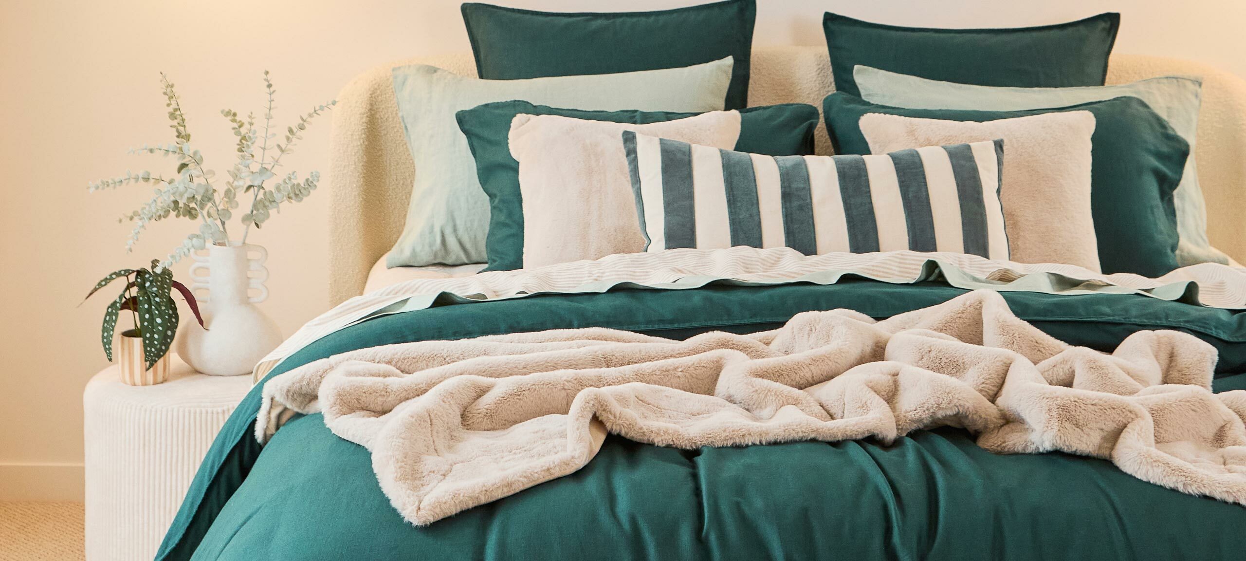 Image features a bed with layers of bedding, including a green quilt cover set, faux fur throw, velvet striped oblong cushion, two square faux fur cushions and green European pillowcases. 