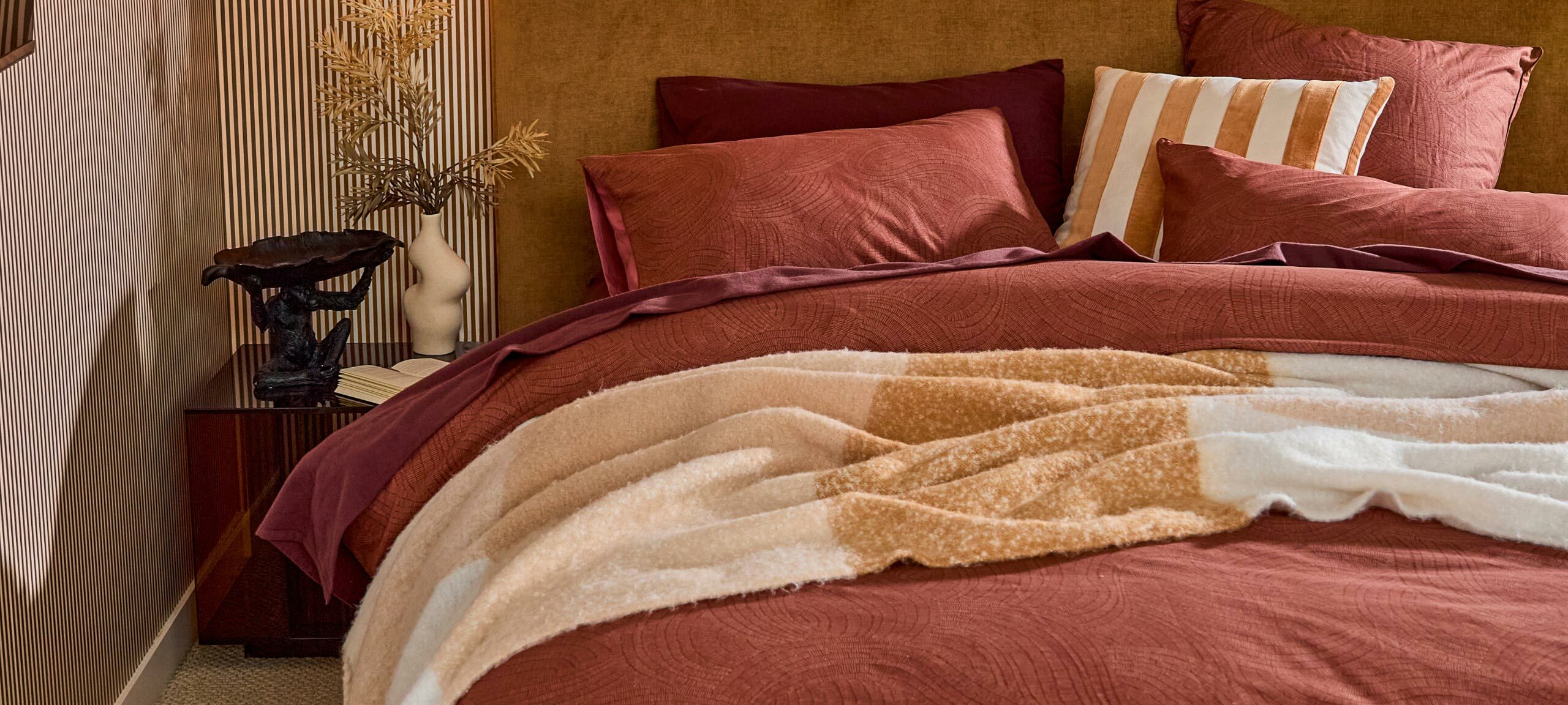 A bed featuring a deep chocolate brown sheet set, rust coloured quilt cover set, a mustard striped cushion and a orange check throw is featured. 