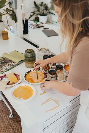 Steph Brooke, a caucasion blonde woman, uses a paint brush to mix colours at her work bench. 
