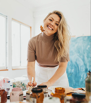 Steph Brooke, a caucasion woman, smiles at the camera while standing at her work bench. Her work bench has paints and other artist tools on it. 