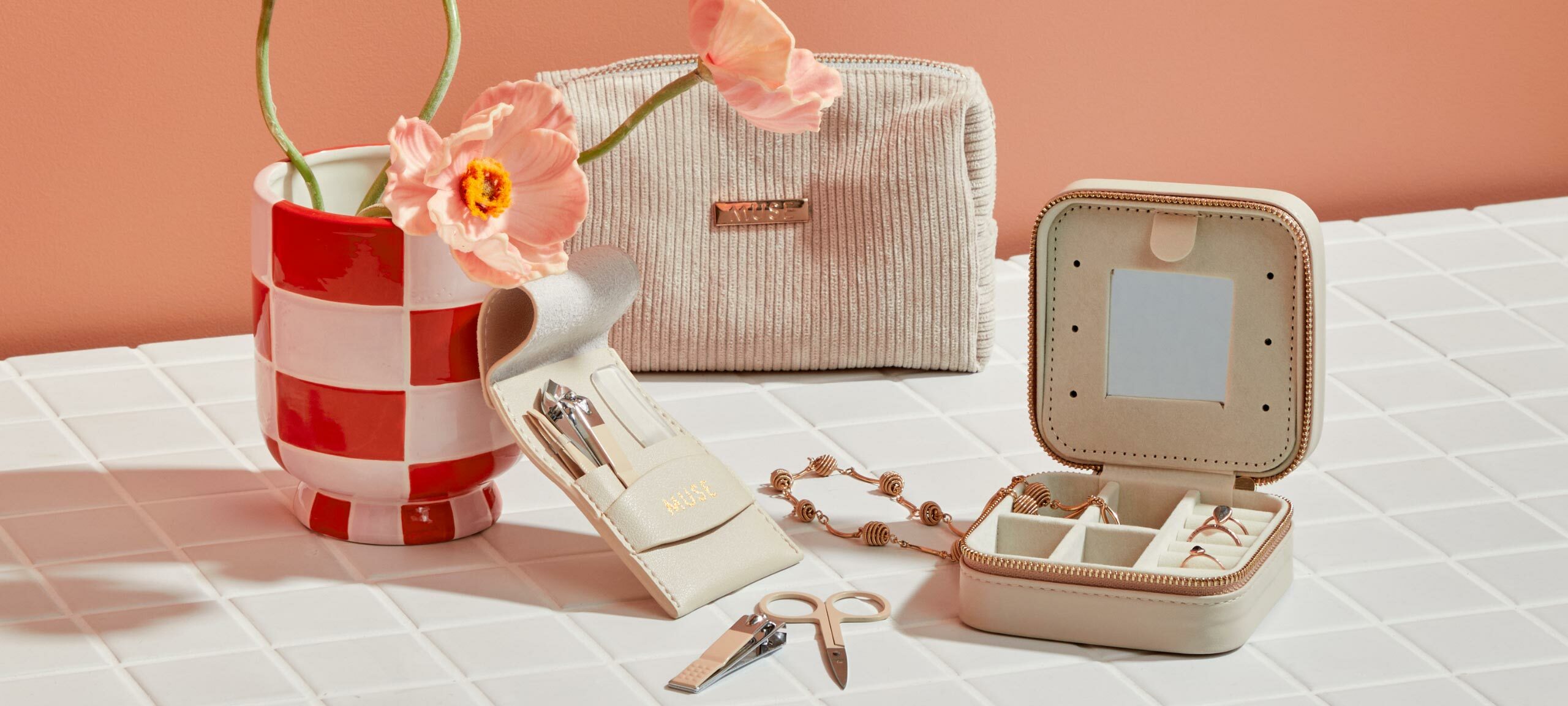 A travel jewellery box, nail care kit including scissors, nail clippers, tweezers and a nail file, a cosmetics bag and a checkered vase sit on top of a tiled counter.  