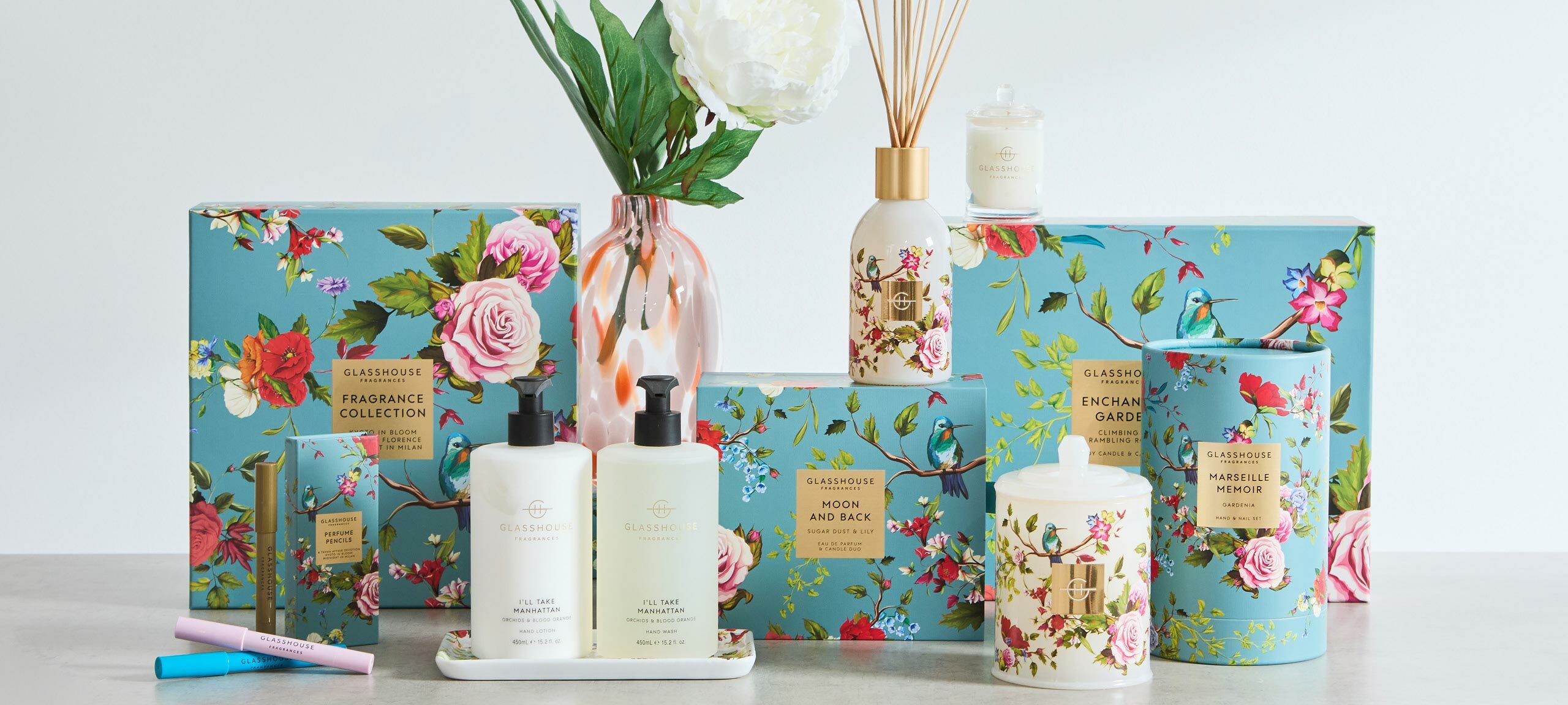 The image pictures Glasshouse's Enchanted Garden gifting range. Each gift box is adorned with a floral print featuring rambling rose vines and a hummingbird. The collection includes a candle, fragrance diffuser, hand care duo, perfume pens and a fragrance collection. 