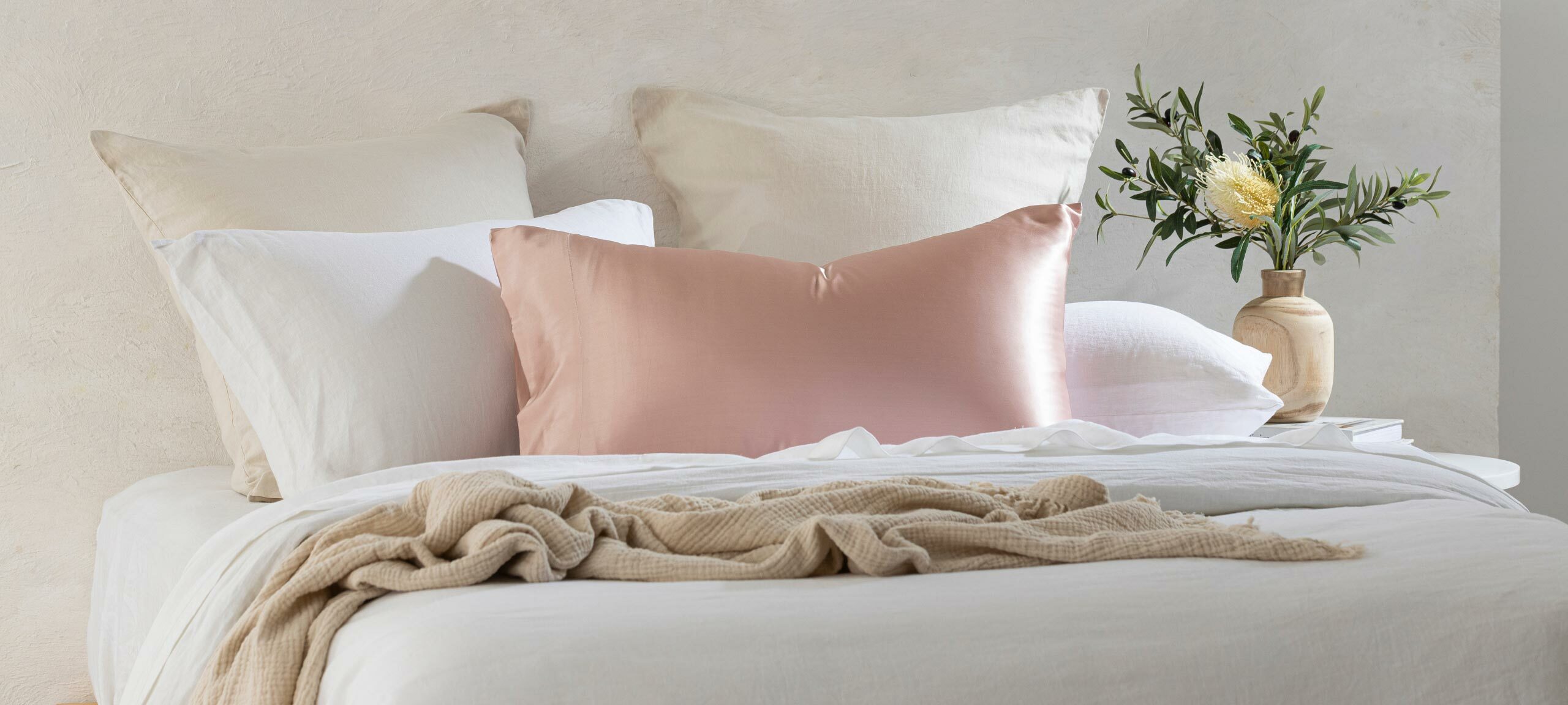 On a bed with white and natural bedding, a pillow sits with a pink silk pillowcase. 