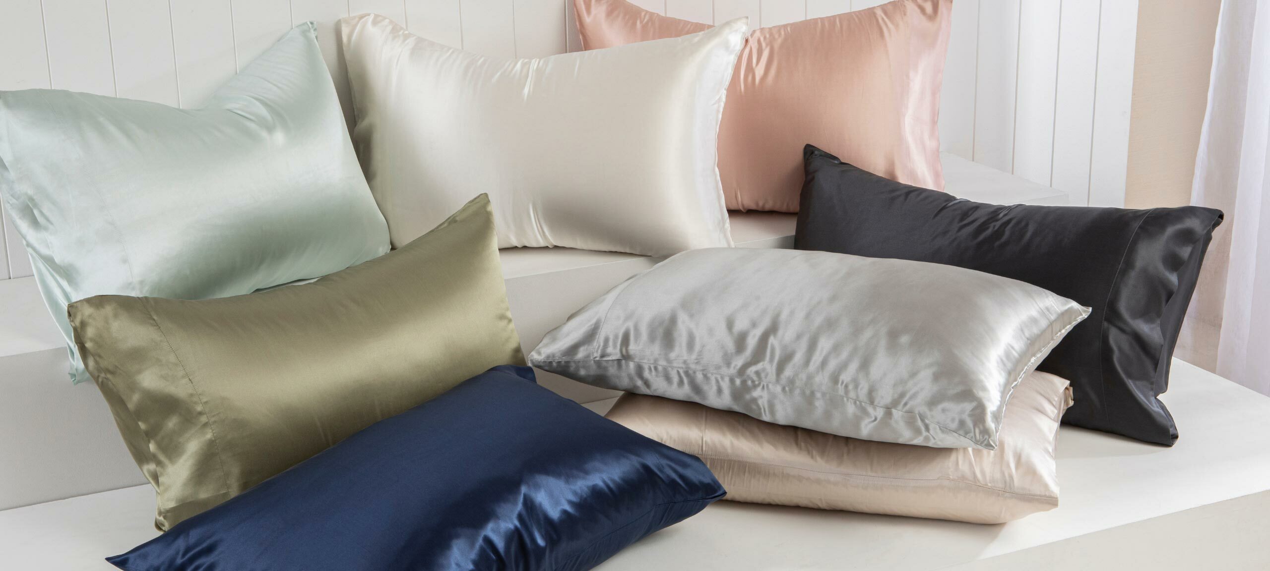 Eight pillows are arranged on a pedestal, each with a different coloured silk pillowcase. From left to right and top to bottom, there is an aqua silk pillowcase, a white silk pillowcase, a pink silk pillowcase, a olive silk pillowcase, a silver silk pillowcase, a black silk pillowcase, a navy blue silk pillowcase and a natural silk pillowcase. 