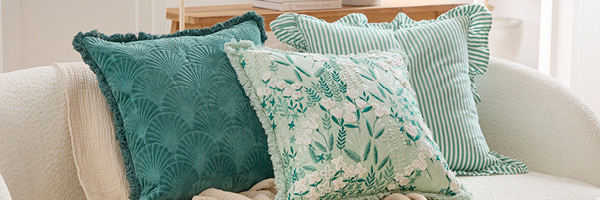 In this picture, three green cushions are styled on a chenille sofa. The hero cushion styled in the front and middle features floral and botanical embroidery and fringing. Behind the hero cushion on the left is a velvet cushion with a quilted fan design and fringed edging. Behind the hero cushion on the right is a striped cushion with a ruffle edge finish. 