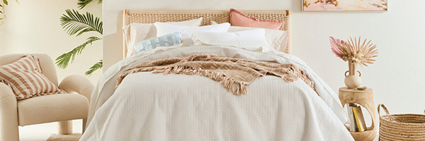 In this picture, a bed with a woven rattan bedhead is styled with a white quilted coverlet, a clay pink cushion, a blue broken check cushion and a rust coloured throw. Beside the bed on the left, a boucle occasional chair is styled with a striped clay and white coloured cushion. On the right, a side table features a vase with dried stems and books in the hollow of the table. 