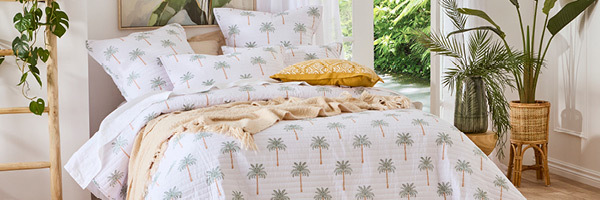 Picture features a bed styled with a coverlet set that features a stylized palm print, a yellow geometric print cushion and a natural coloured throw. Around the bed there are artificial plants styled in vases and planters. 