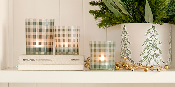 Three gingham candle holders with candles in them sit on a shelf