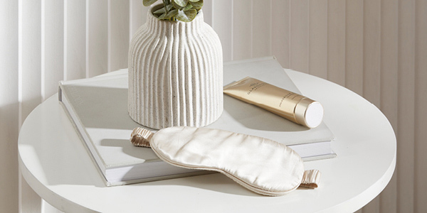 A silk eye mask, hand lotion and decorative artificial plant sit on a bedside table