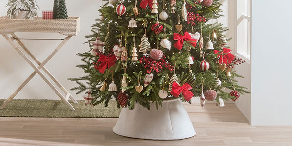 A Christmas tree decorated with ornaments, bows, holly stems, lights and a white tree skirt. 